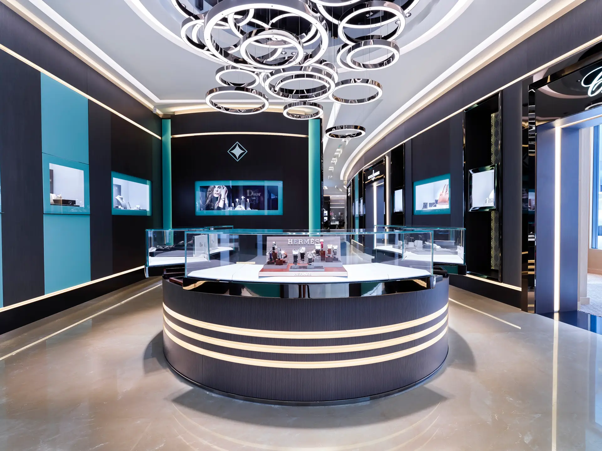Internationales Luxus-Lichtdesign von OZ Lighting im High-End Flagship-Showroom von Ahmed Seddiqi & Sons in Dubai. Mehrere ineinander verschlungene, kreisförmige LED-Ringleuchten an der Decke kombinieren sich mit präzisen Einbauspots, um exklusive Uhren- und Schmuckvitrinen von Hermes und Chopard reflexionsfrei und brillant auszuleuchten.