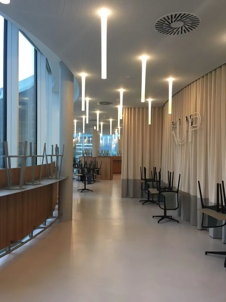 Moderne Gastronomie-Beleuchtung von OZ Lighting im Foodcourt des DC Tower in Wien. Vertikal abgehängte, linienförmige LED-Pendelrohre an einer perforierten Akustikdecke setzen dynamische Lichtakzente im Korridor und entlang der hölzernen Counter-Bereiche, kombiniert mit minimalistischem Interior Design und kontrastierenden Lichtstrukturen für eine urbane Food-Mall-Atmosphäre.