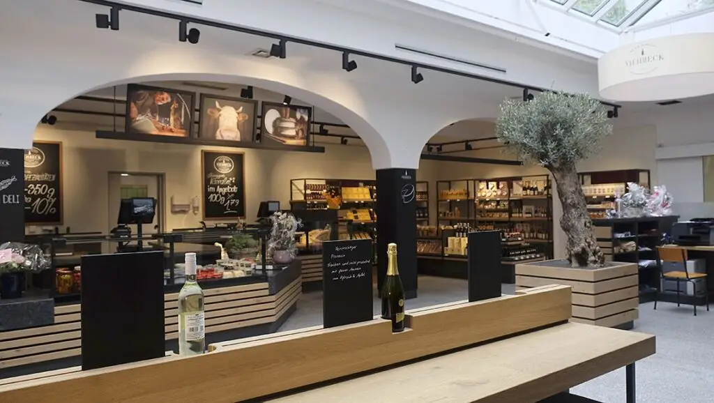 Ganzheitliche Lichtarchitektur von OZ Lighting im Delikatessengeschäft Viehbeck. Die Symbiose aus natürlichem Oberlicht, architektonischen weißen Rundbögen und abgehängten, schwarzen LED-Stromschienen-Spots über den Holztheken sorgt für eine einladende Marktatmosphäre.