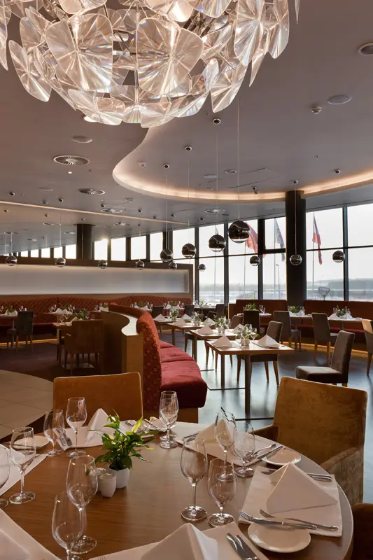 Luxuriöse Hotel- und Restaurantbeleuchtung im Crowne Plaza von OZ Lighting. Ein skulpturaler Glas-Chandelier bricht das Licht kunstvoll, während eine geschwungene, warmweiße Decken-Voute (indirektes Licht) und metallische Pendelspots den Raum strukturieren.
