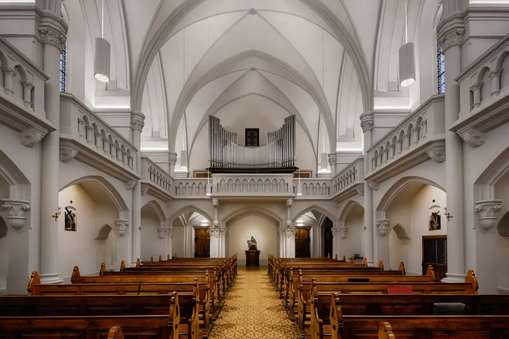 Sakrale Architekturbeleuchtung und Lichtplanung von OZ Lighting in der Kirche der Kreuzschwestern in Graz. Minimalistische, zylindrische weiße LED-Pendelleuchten sorgen für eine harmonische Grundbeleuchtung des Kirchenschiffs und der Holzbank-Reihen, während verdeckt installierte, warmweiße LED-Lichtbänder die neogotischen Deckengewölbe, Emporen und den Orgelprospekt indirekt illuminieren.