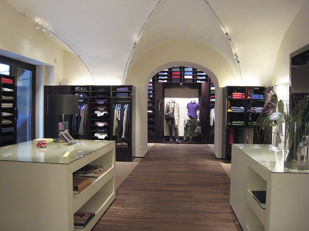 Exklusive Ladenbeleuchtung und Lichtplanung von OZ Lighting in der Mode Grüner Fashion-Boutique. Ein filigranes, geschwungenes LED-Schienensystem folgt der Architektur des historischen Kreuzgewölbes und setzt Verkaufsflächen, Kleiderständer und Mannequins durch fokussierte Akzentbeleuchtung perfekt in Szene, ergänzt durch indirektes Uplighting zur Betonung der Deckenstruktur.