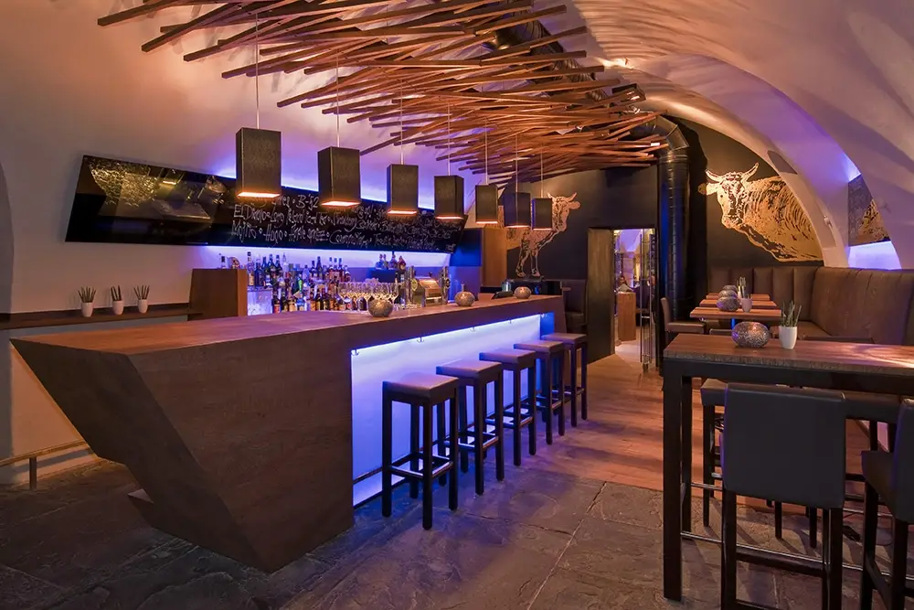 Stimmungsvolle Gastronomie-Beleuchtung im Steak-Restaurant el Gaucho umgesetzt von OZ Lighting. Eine Bar mit tiefblauer LED-Thekenbeleuchtung harmoniert mit einer Deckenkonstruktion aus Altholz und minimalistischen, kubischen Pendelleuchten für ein exklusives Ambiente.