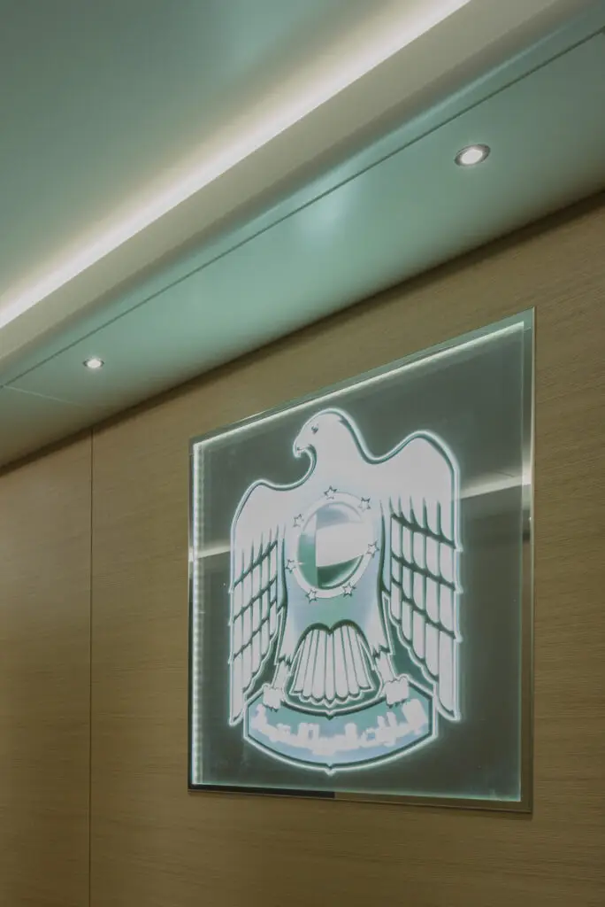 Detailaufnahme eines kanteneingespeisten LED-Acryl-Lichtschilds von OZ Lighting im Schabus VIP-Container. Das präzise eingravierte offizielle Wappen der Vereinigten Arabischen Emirate (VAE) mit dem ikonischen Falken-Emblem wird durch moderne LED-Effektbeleuchtung illuminiert und von bündigen Decken-Spots flankiert.
